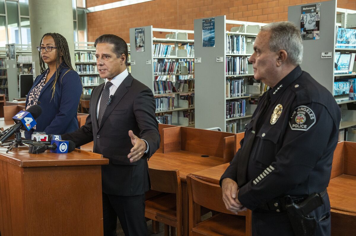 A man in a suit speaks at a lectern in a school library, flanked by a uniformed school police official and a woman in a suit