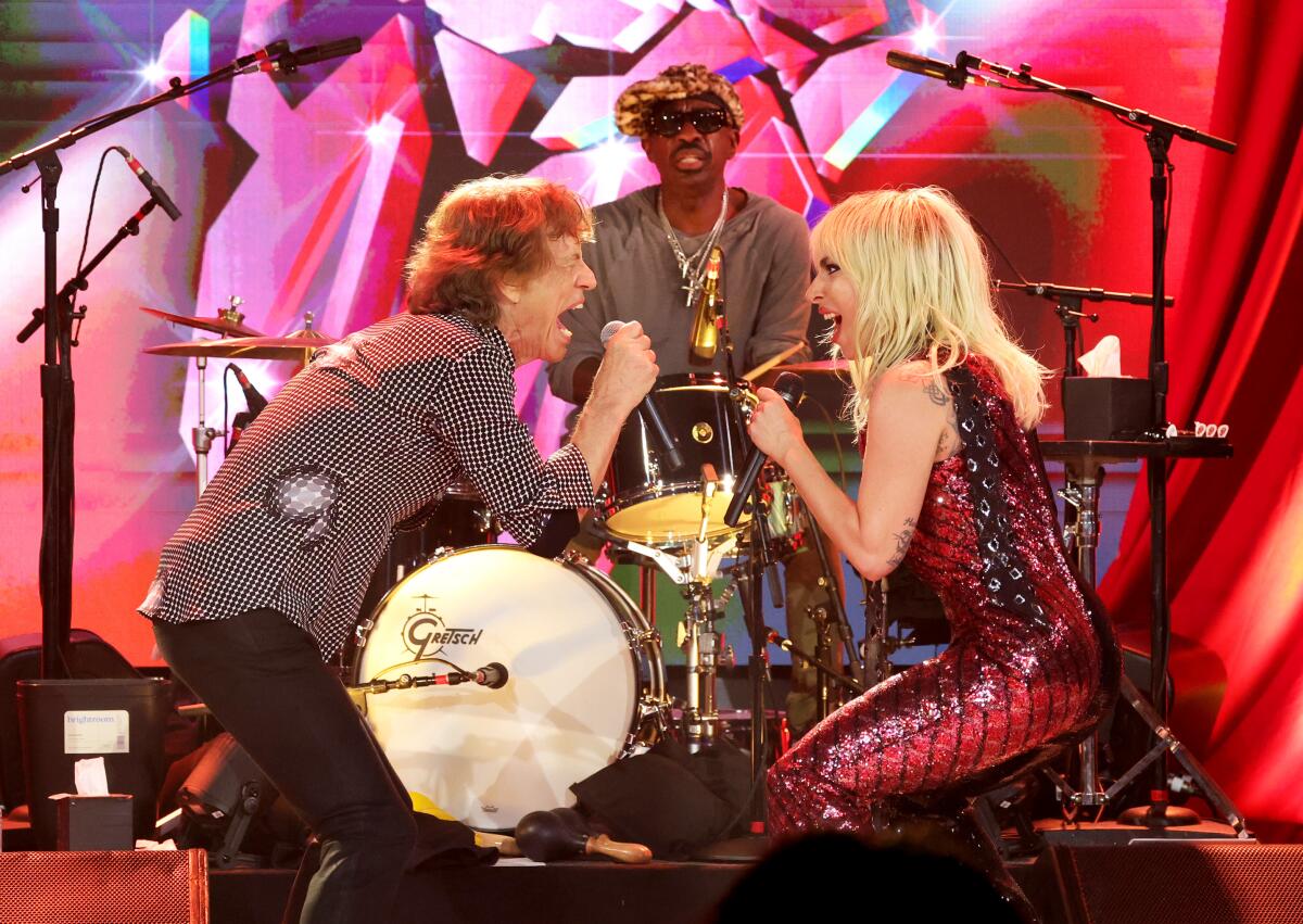 Mick Jagger and Lady Gaga hold microphones and face each other as they sing on a stage in front of a drummer