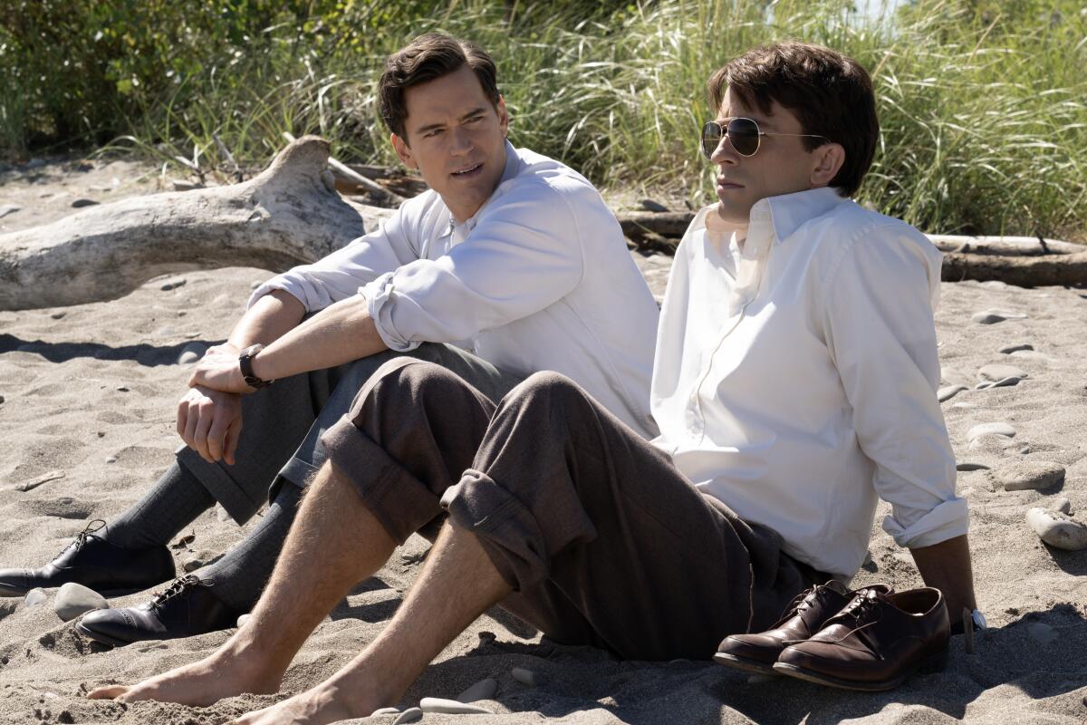 Matt Bomer and Jonathan Bailey, in white shirts and khakis, sit on a beach.