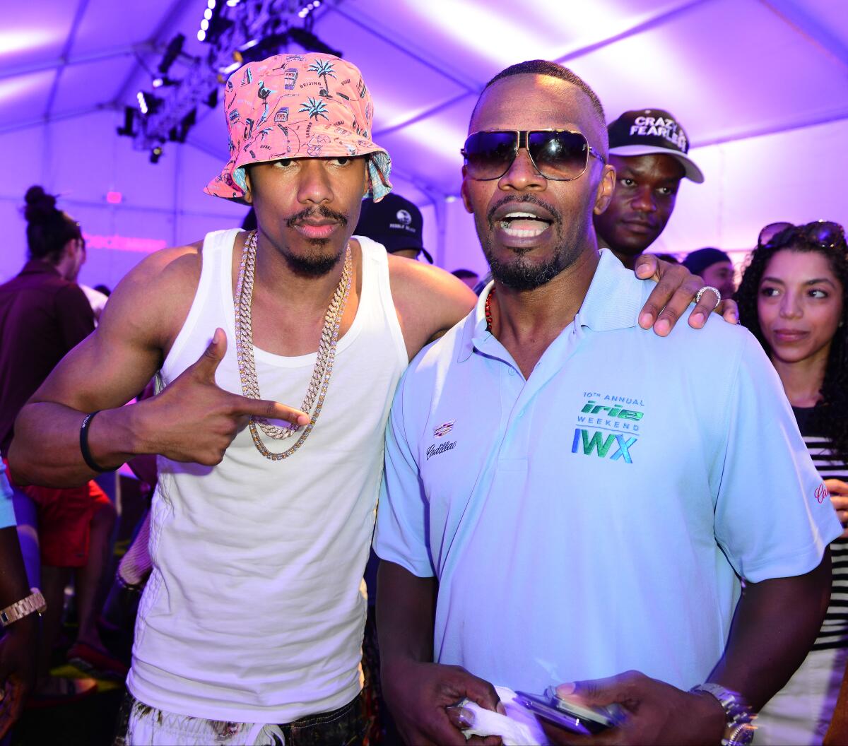 Nick Cannon, wearing a bucket hat and white tank top, poses next to Jamie Foxx, who is in a blue polo shirt and sunglasses