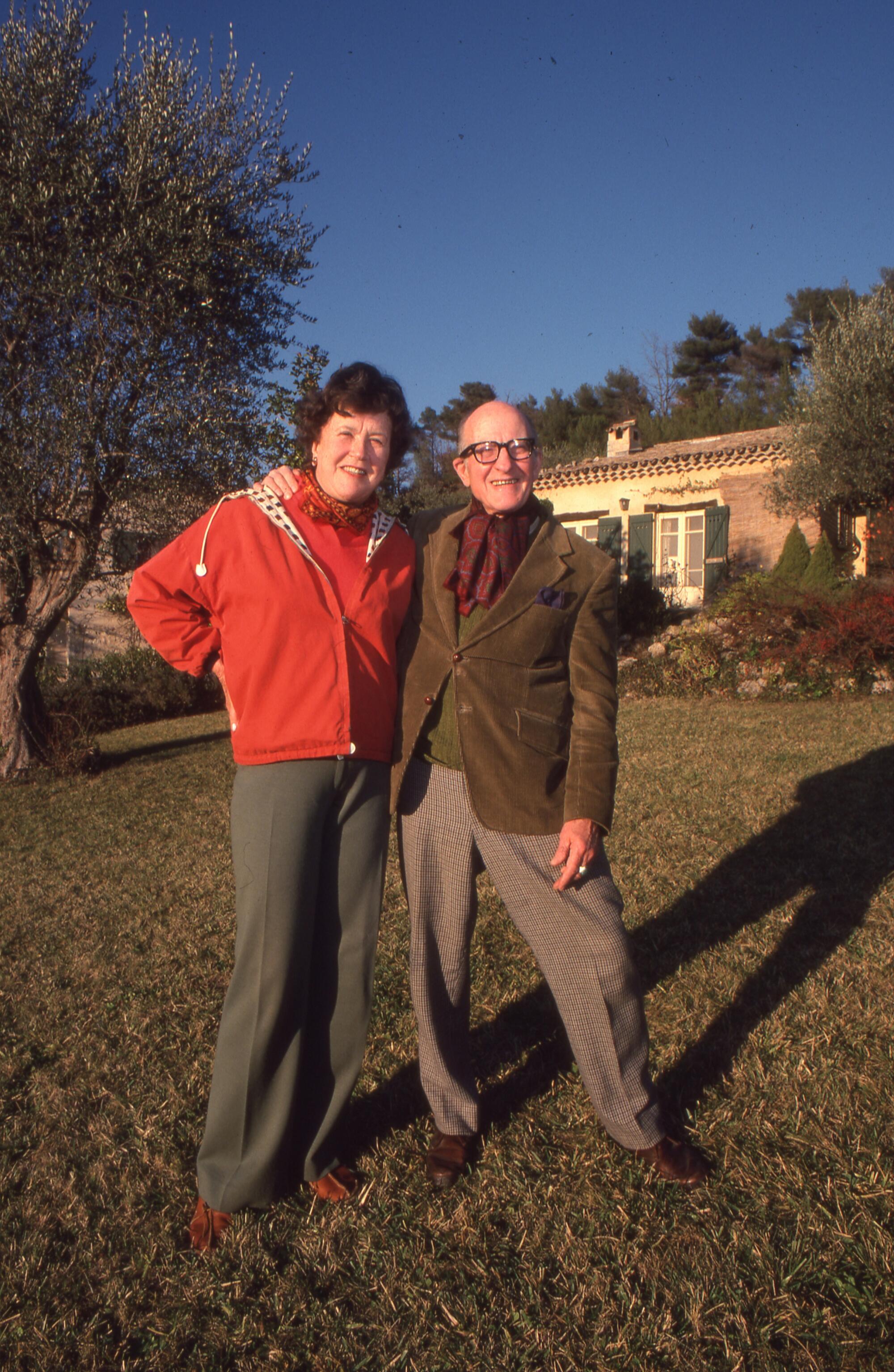 Julia Child n in a red sweater and Paul Child in an olive jacket with their arms around each other outside on a nice day 