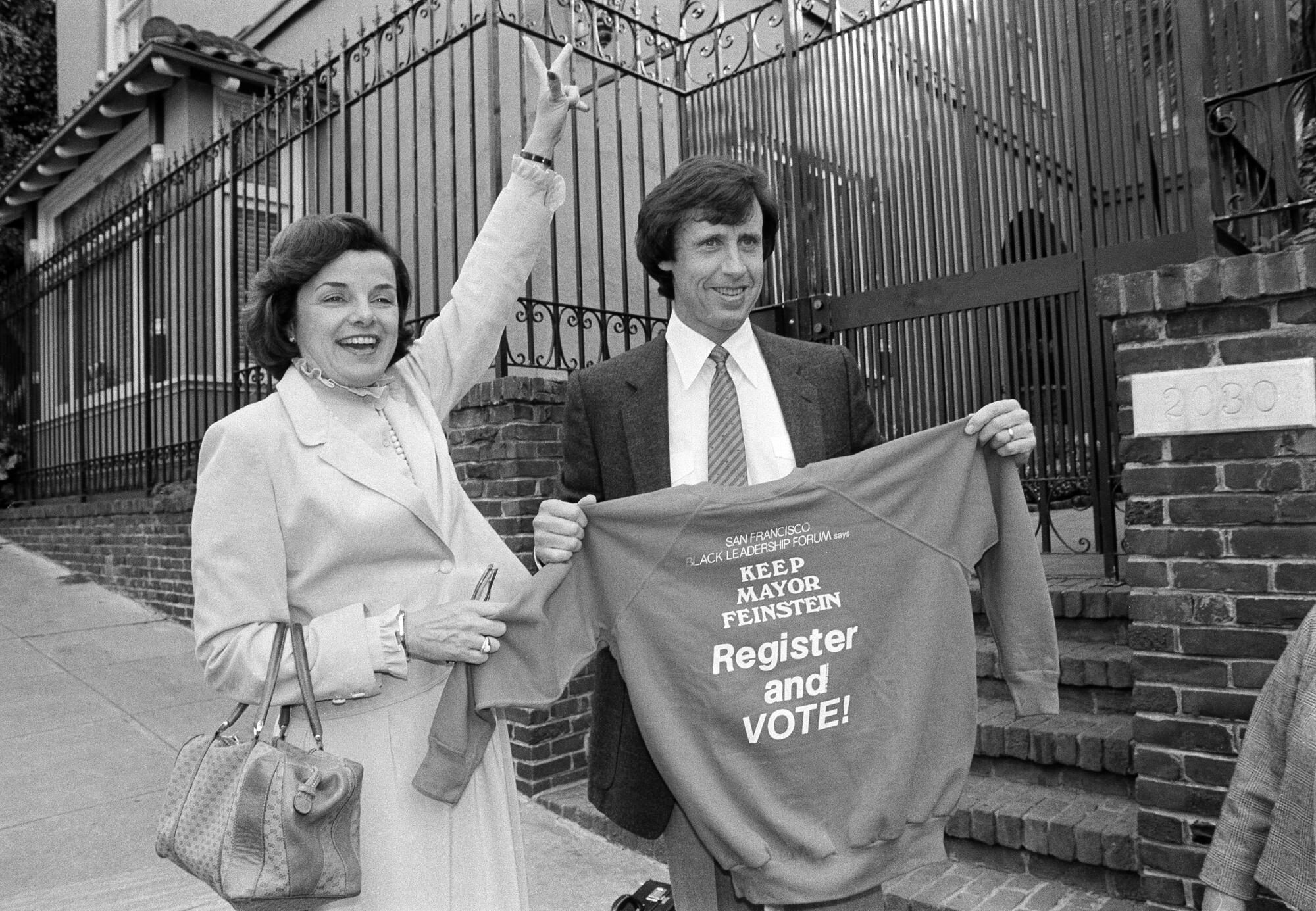 A black-and-white photo of San Francisco Mayor Dianne Feinstein flashing a V sign as Richard Blum holds up a campaign shirt