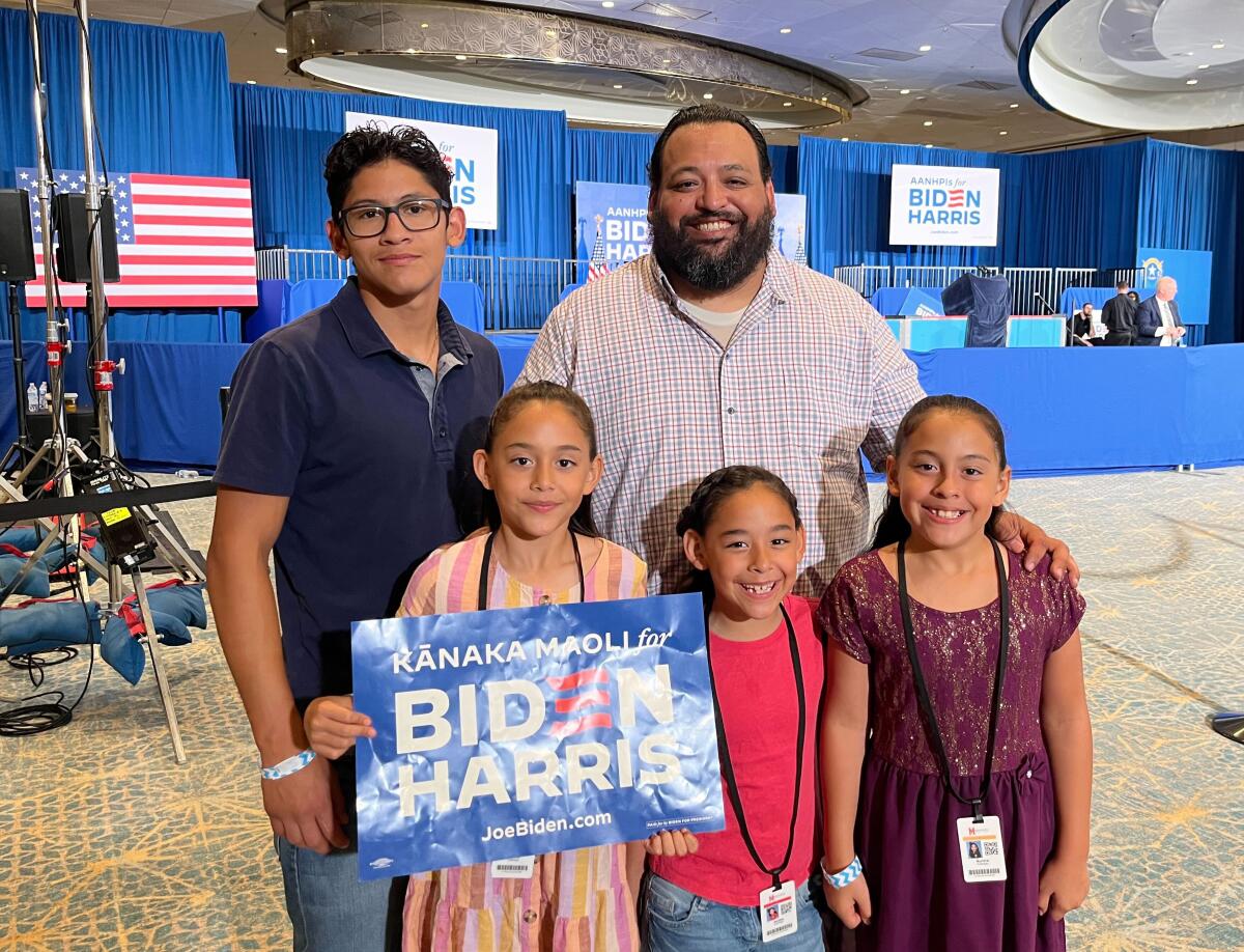 A man standing with a younger man behind three young girls, one holding a blue Biden-Harris sign, in front of a blue stage