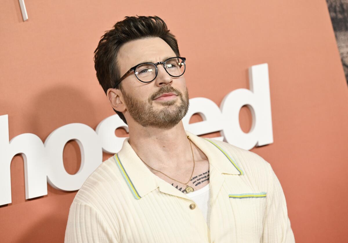 Chris Evans poses in glasses and a cream collared shirt against an orange background.