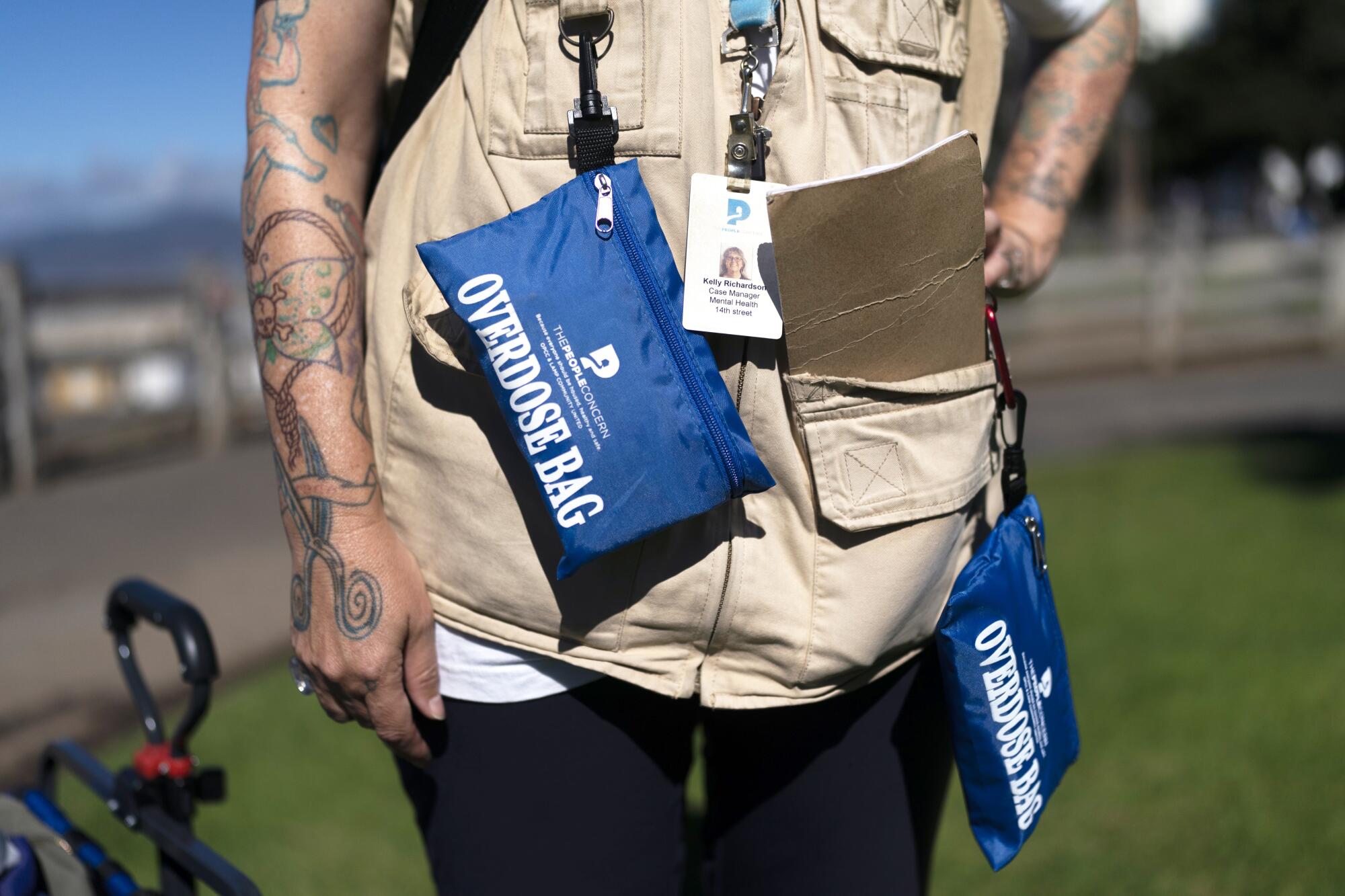 A torso view of a person wearing a light-brown west with small bags hanging off of it that read "Overdose Bag."