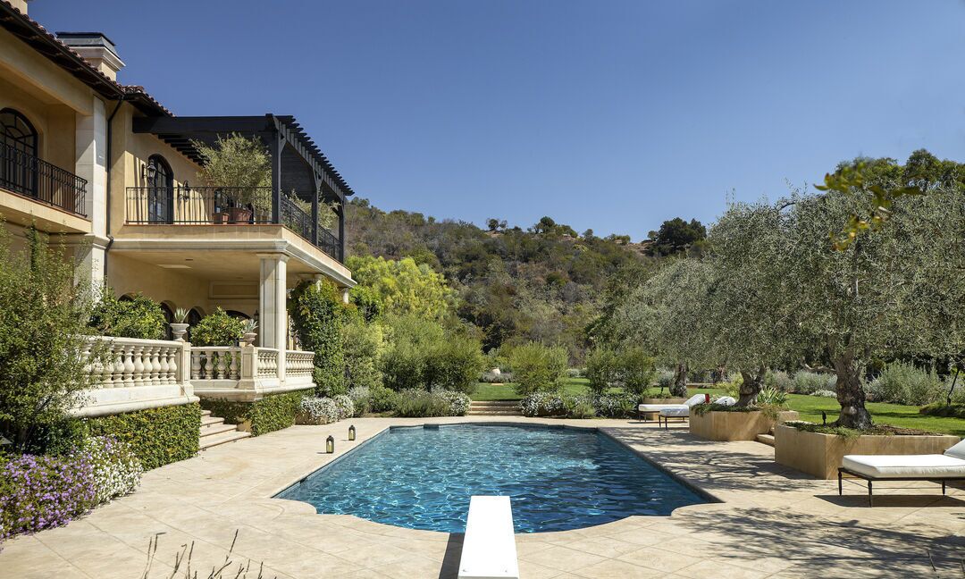 The pool has a diving board and it sits in front of the house amid trees.