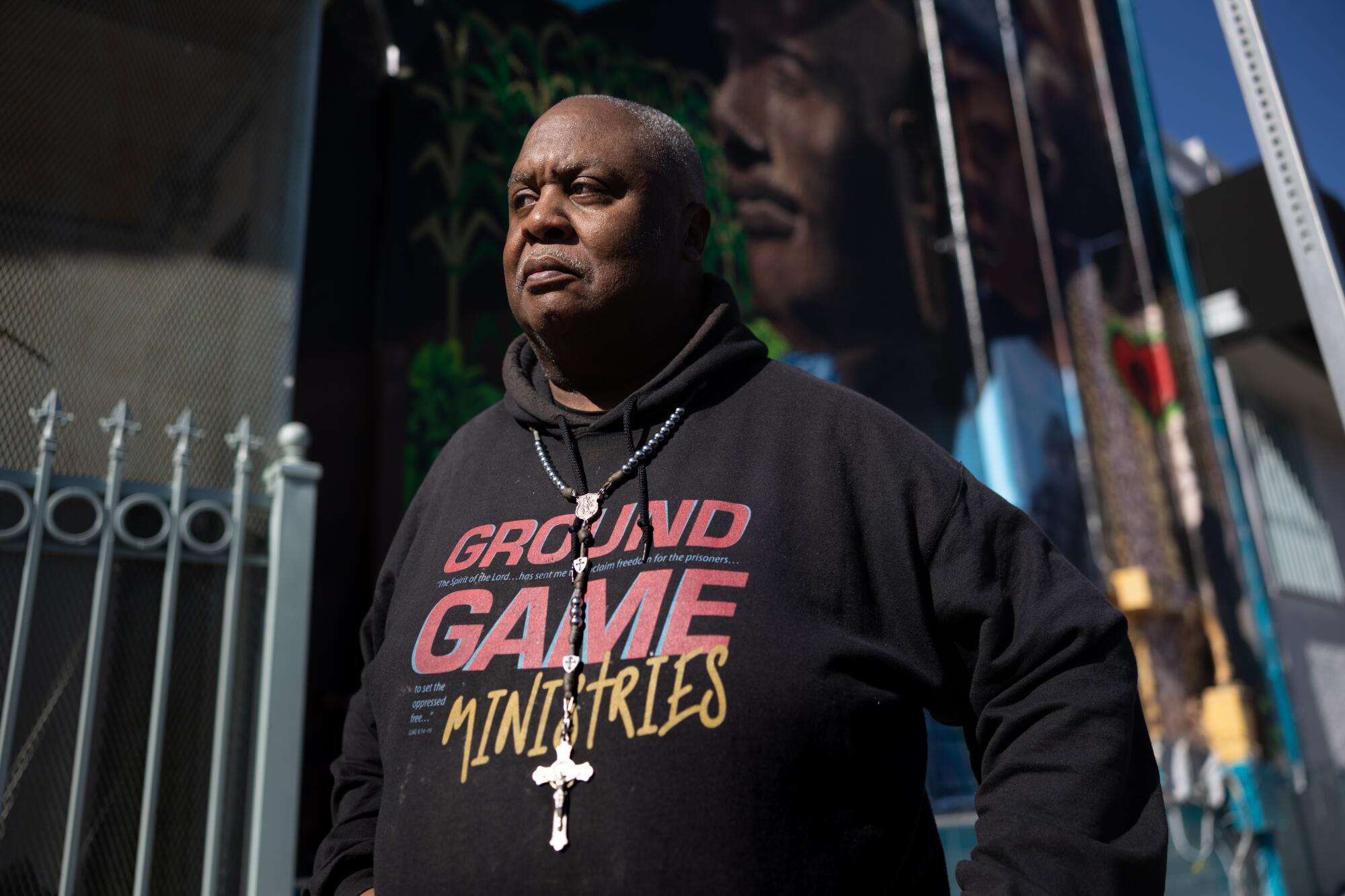 A man wearing a cross and a black hoodie reading "Ground Game Ministries" stands front of a mural after dark