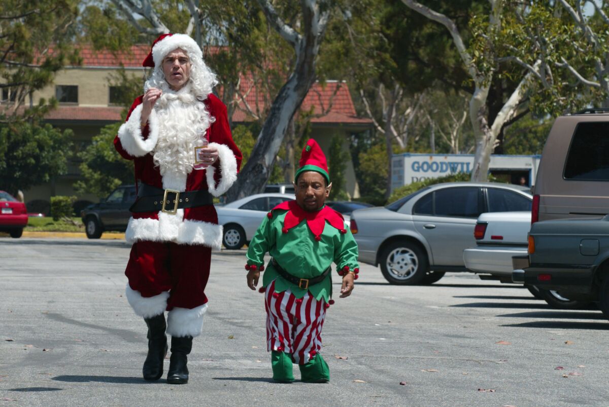A tall white man dressed as Santa Claus and a Black little person in an elf costume stroll through a parking lot.