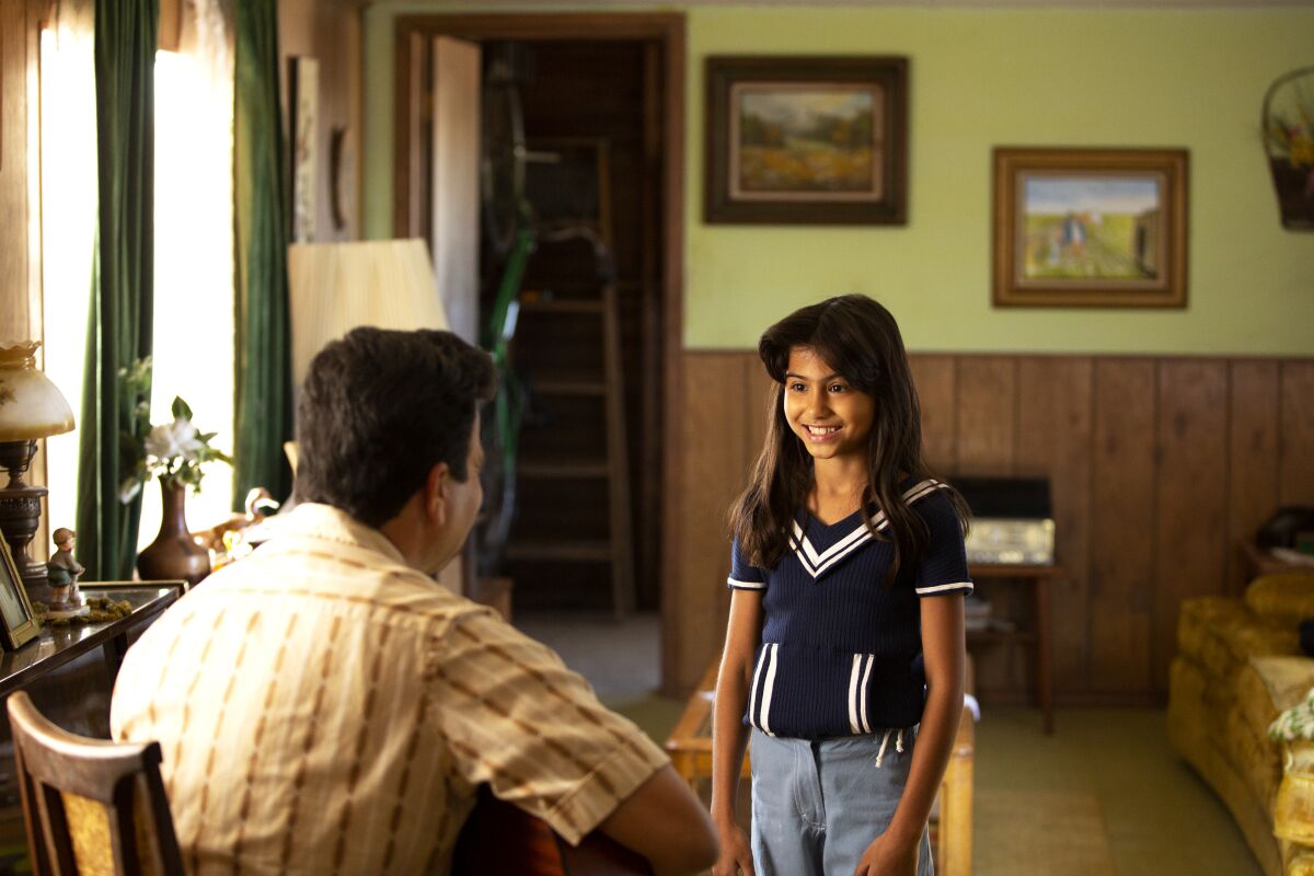 Young Selena, played by Madison Taylor Baez, smiles a gap-toothed grin at her father, seated and playing guitar.