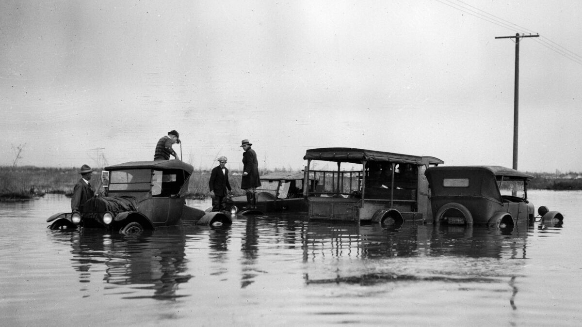 Four people stand on top of 1920s cars in a flooded intersection