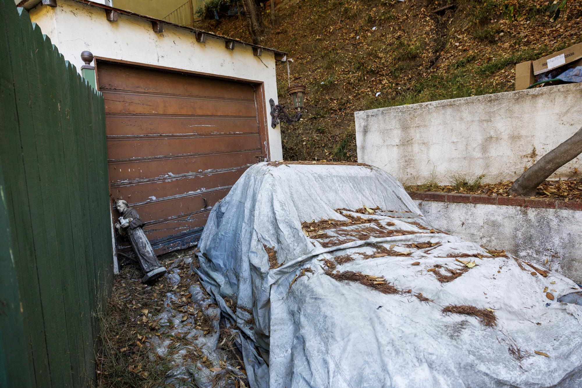 A vehicle under a white cover in front of a garage door, leaves and evergreen needles scattered on the car and driveway