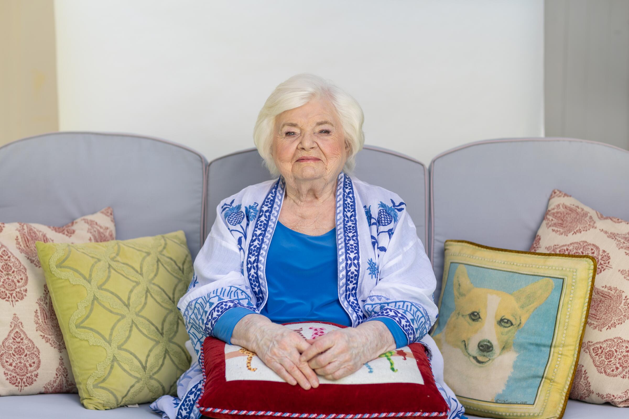 A smiling elderly woman sits on a couch next to her dog pillow.