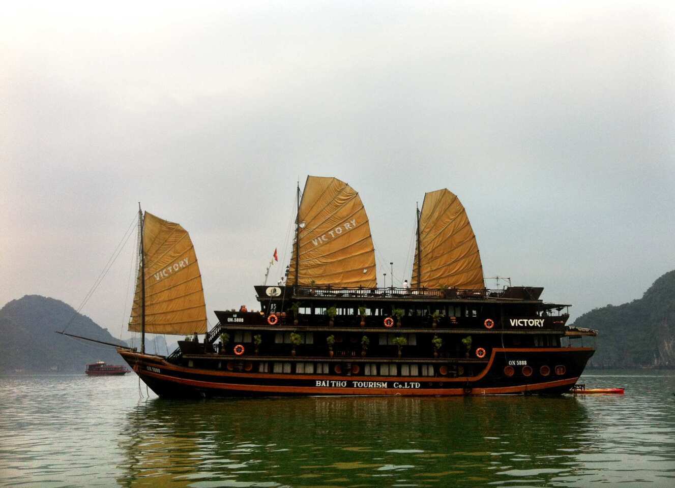 A junk cruise ship raises its eye-catching sails as it plies the emerald waters of Halong Bay, Vietnam. A kayak roped to the back of the ship can be rented to paddle to a little islet filled with monkeys.