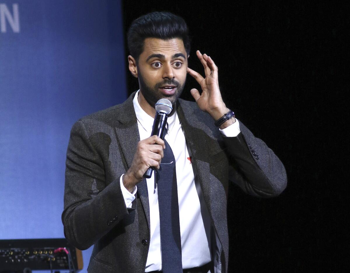 Hasan Minhaj wears a dark suit and tie while speaking into a handheld microphone onstage