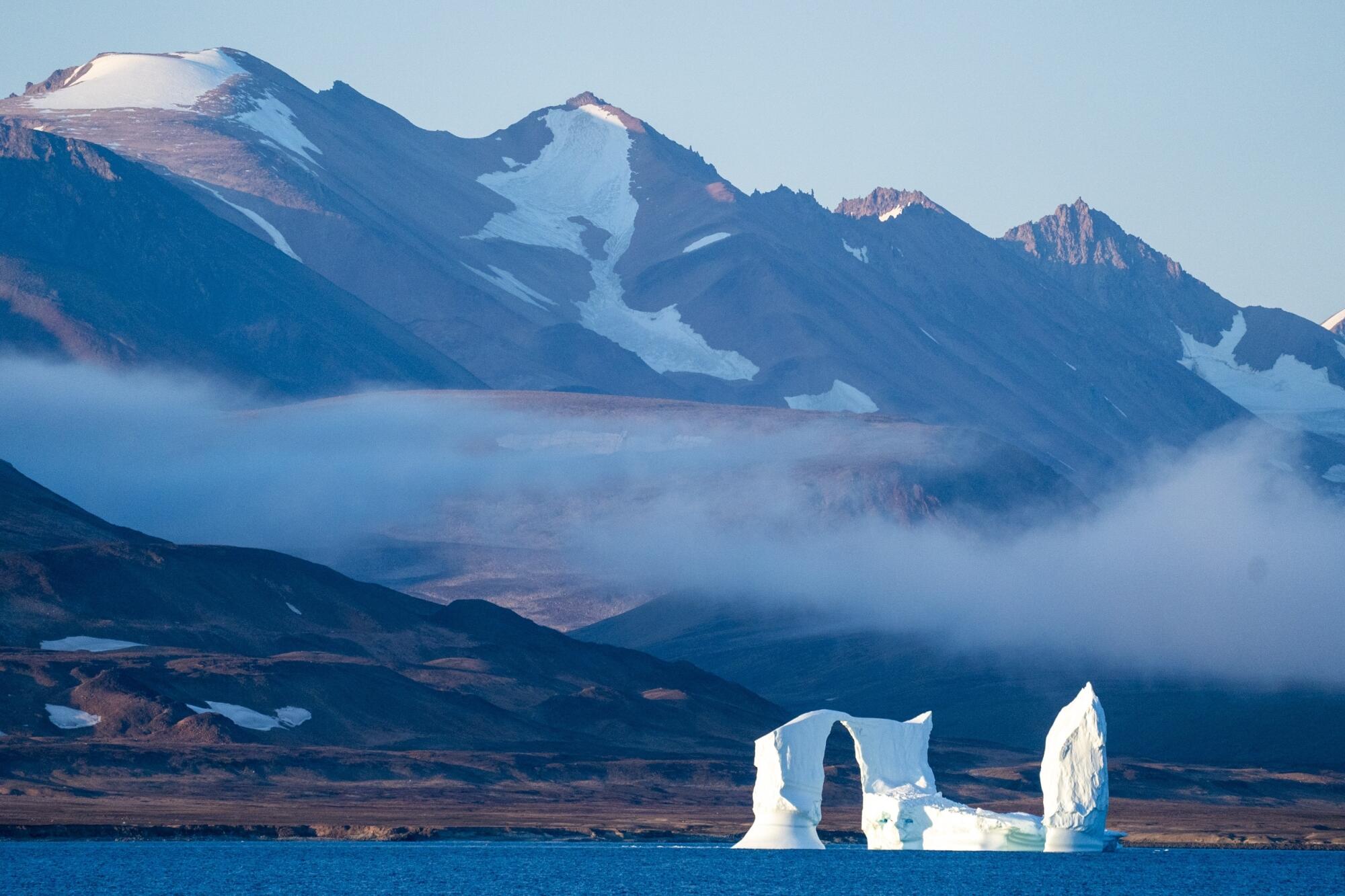 An iceberg floats in the sea as mountains rise in the background.