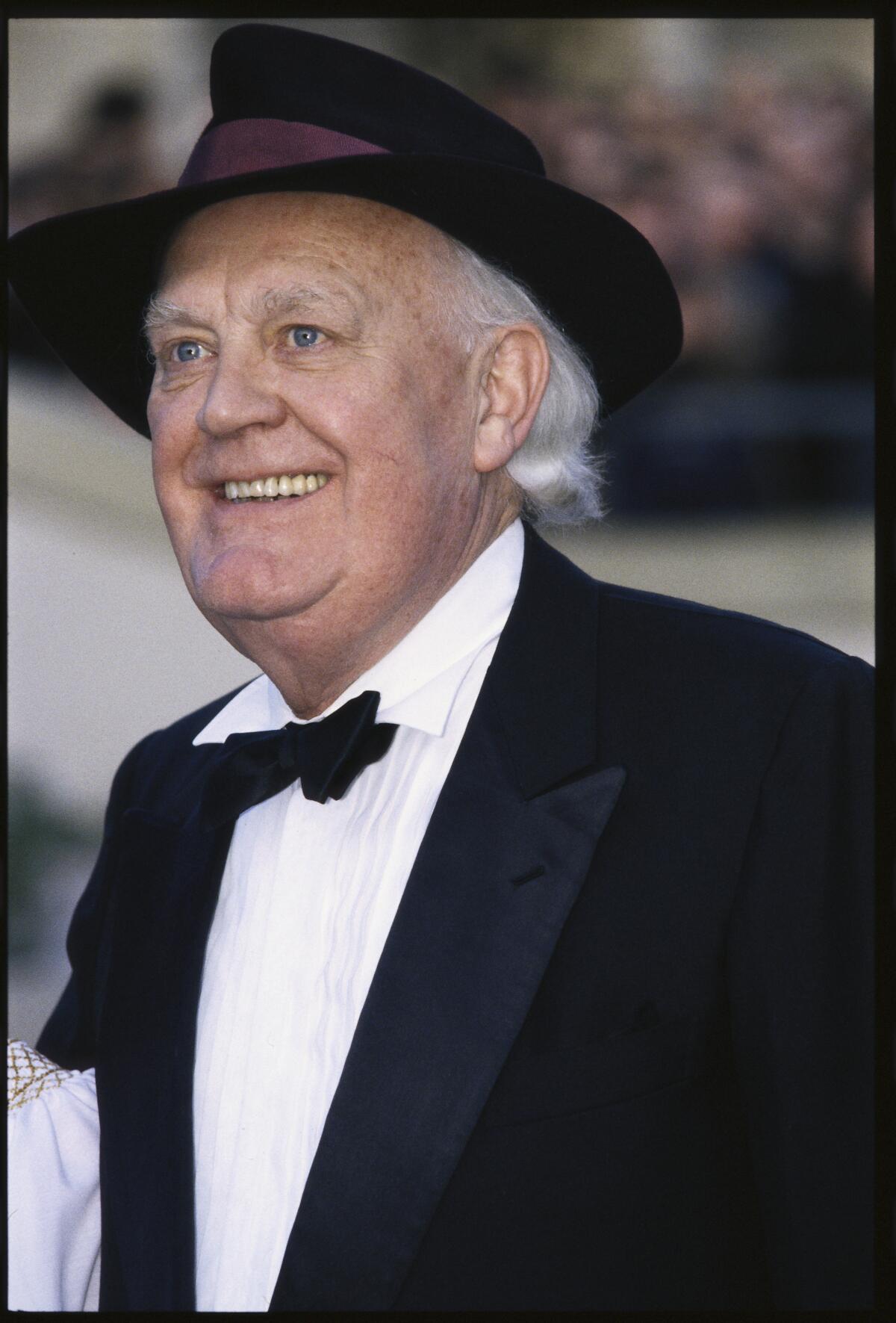 Joss Ackland wears a black tuxedo with a black top hat as he poses for pictures while on a red carpet.