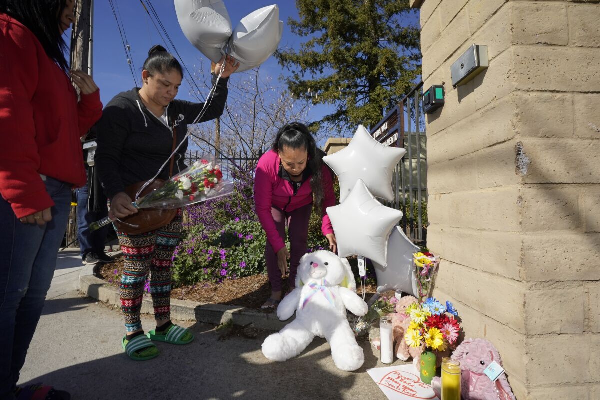 A woman brings flowers and balloons to a memorial while another places a large white teddy bear near candles and other items.