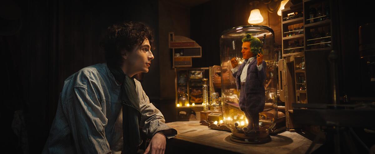 Timothée Chalamet as Willy Wonka looks into a glass jar containing an Oompa Loompa played by Hugh Grant