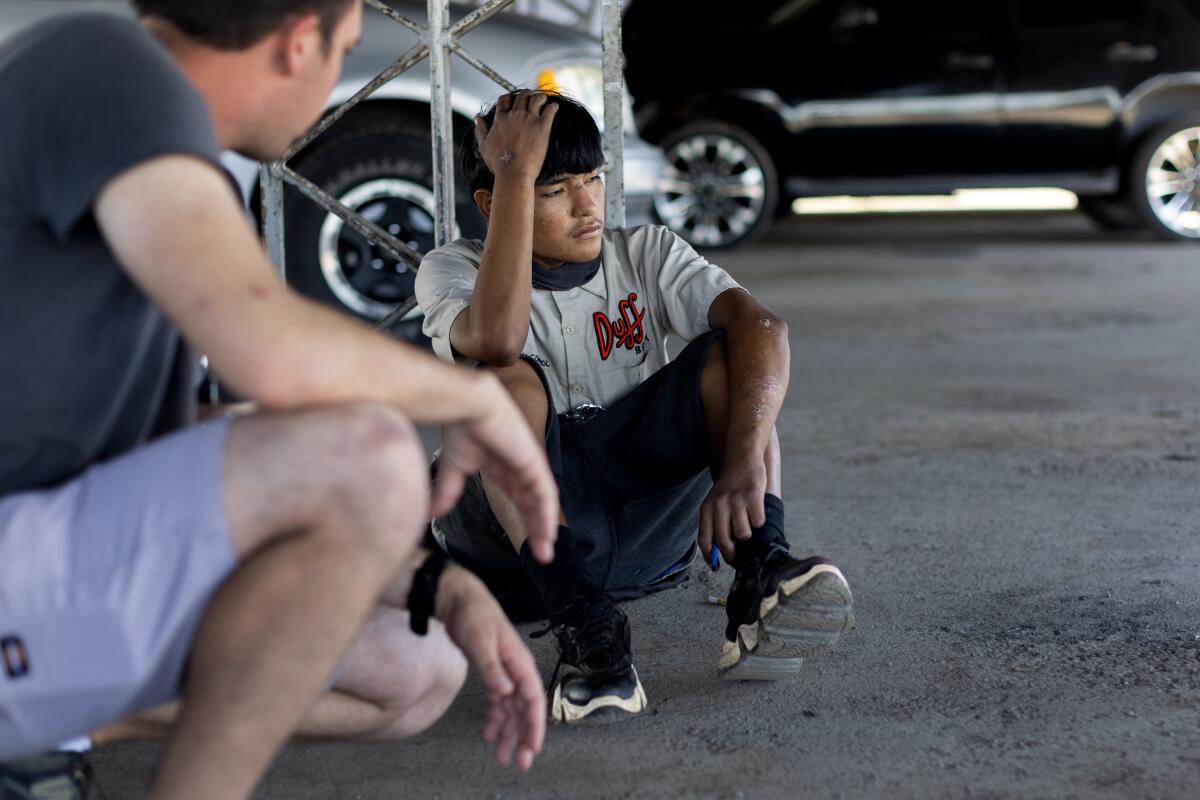 A young man sitting on pavement, a hand on his head, looking away as another man crouches down to talk with him