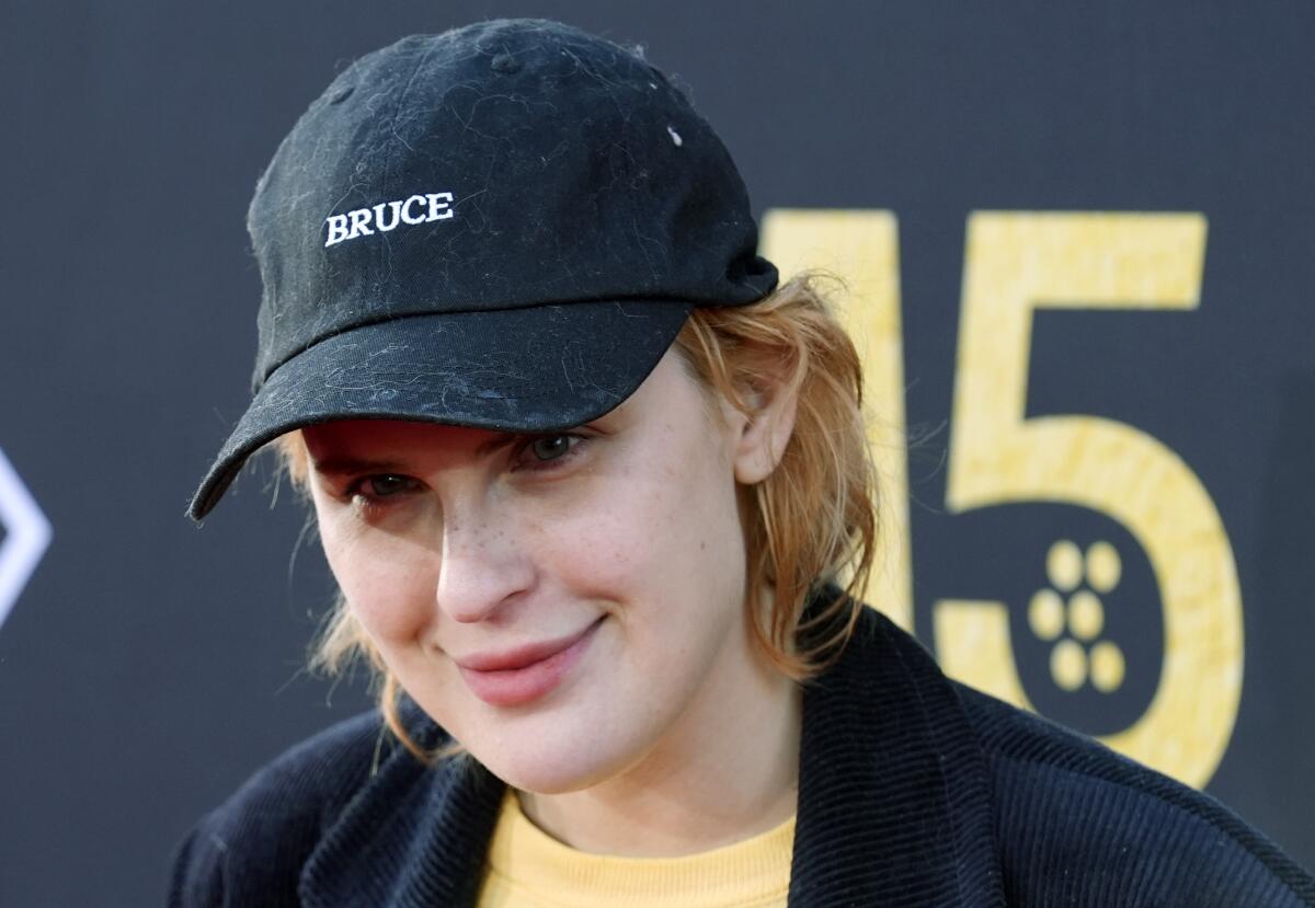 Tallulah Willis smirking, wearing a dark hat with the word 'Bruce' on it. 