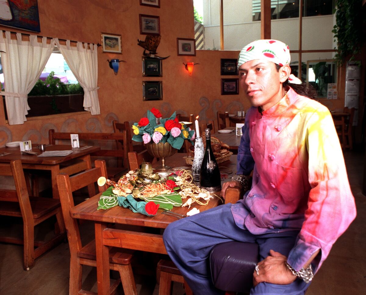  A man in a colorful chef's uniform, a kerchief covering his hair, sits at a table with food and wine