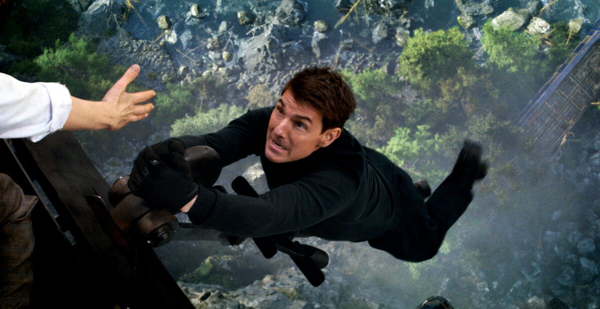 A hand reaches out to help Tom Cruise hanging onto the wheel of an ascending aircraft