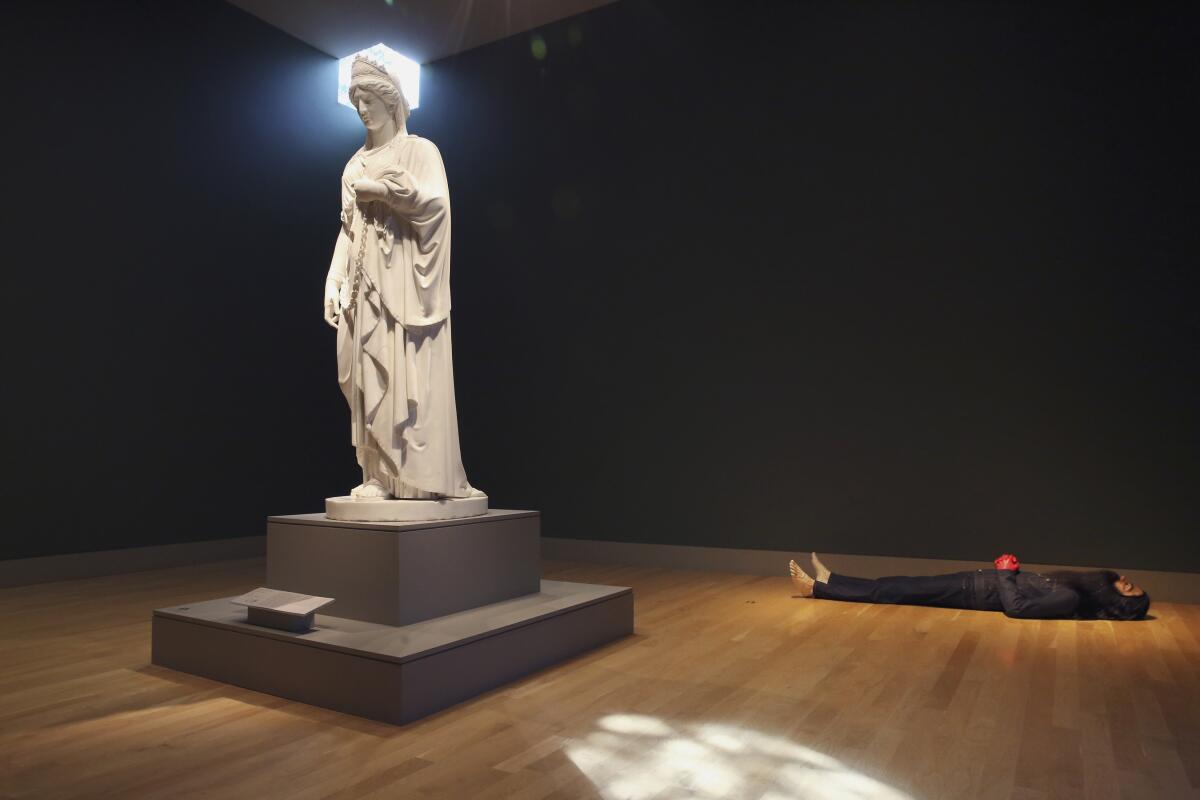 A marble figure stands next to a contemporary sculpture of a prone person