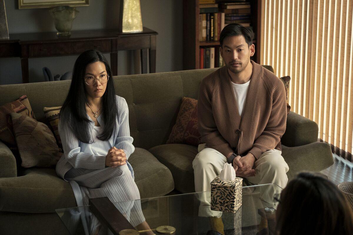 A woman and a man sit on a couch, each with their hands folded in their lap.