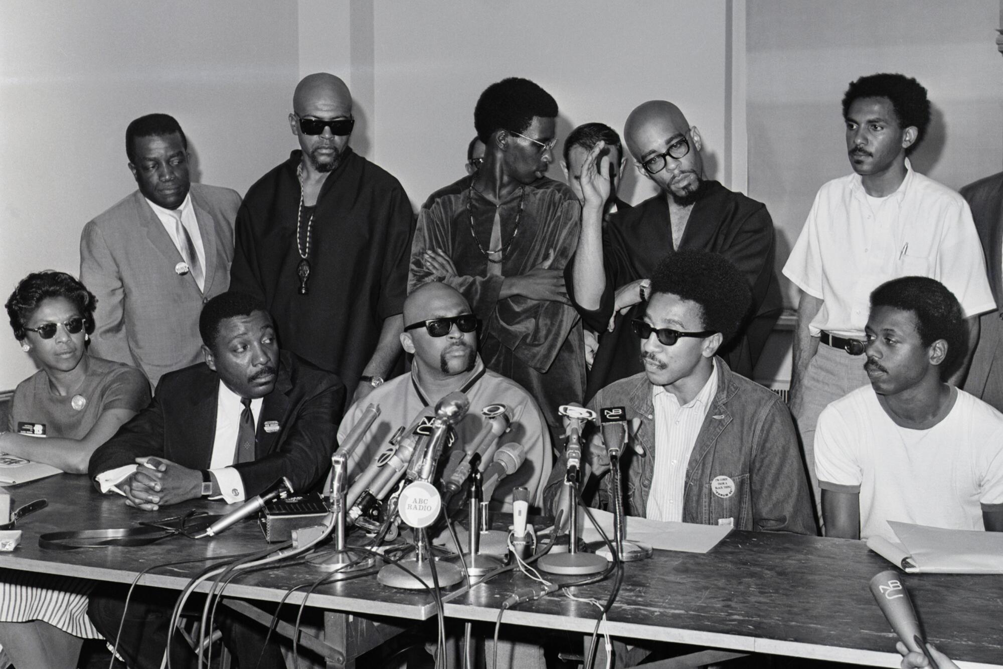 A woman is seated at far left alongside four men at microphones, and several more men standing behind