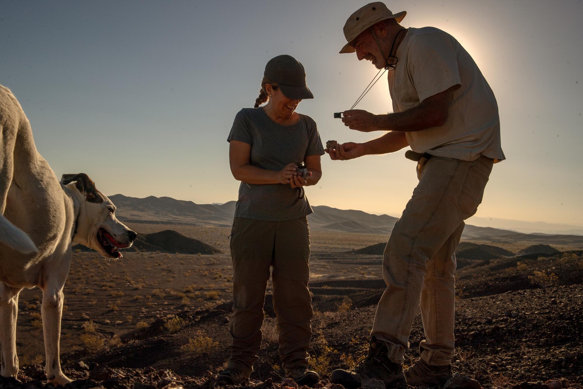 A man and a woman examine a rock sample in a desert landscape as a dog looks on.