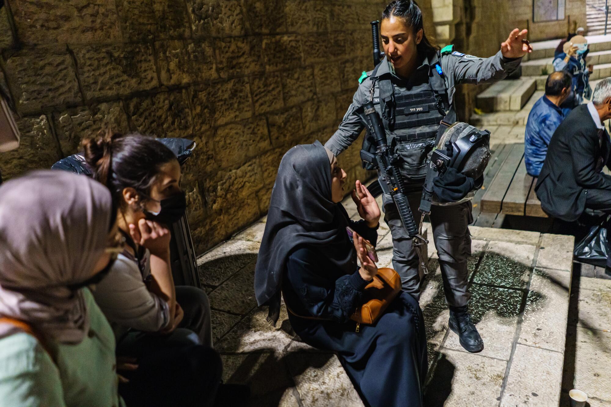 A young woman in military uniform nudges a seated woman in a hajib, who holds up both hands.