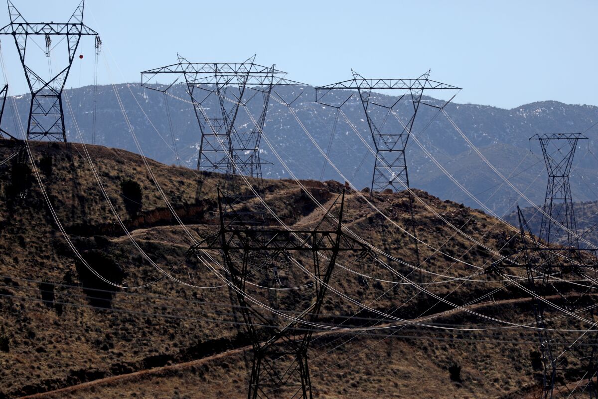 A row of electric transmission lines run on a ridge of a dry hill, with mountains on the horizon.