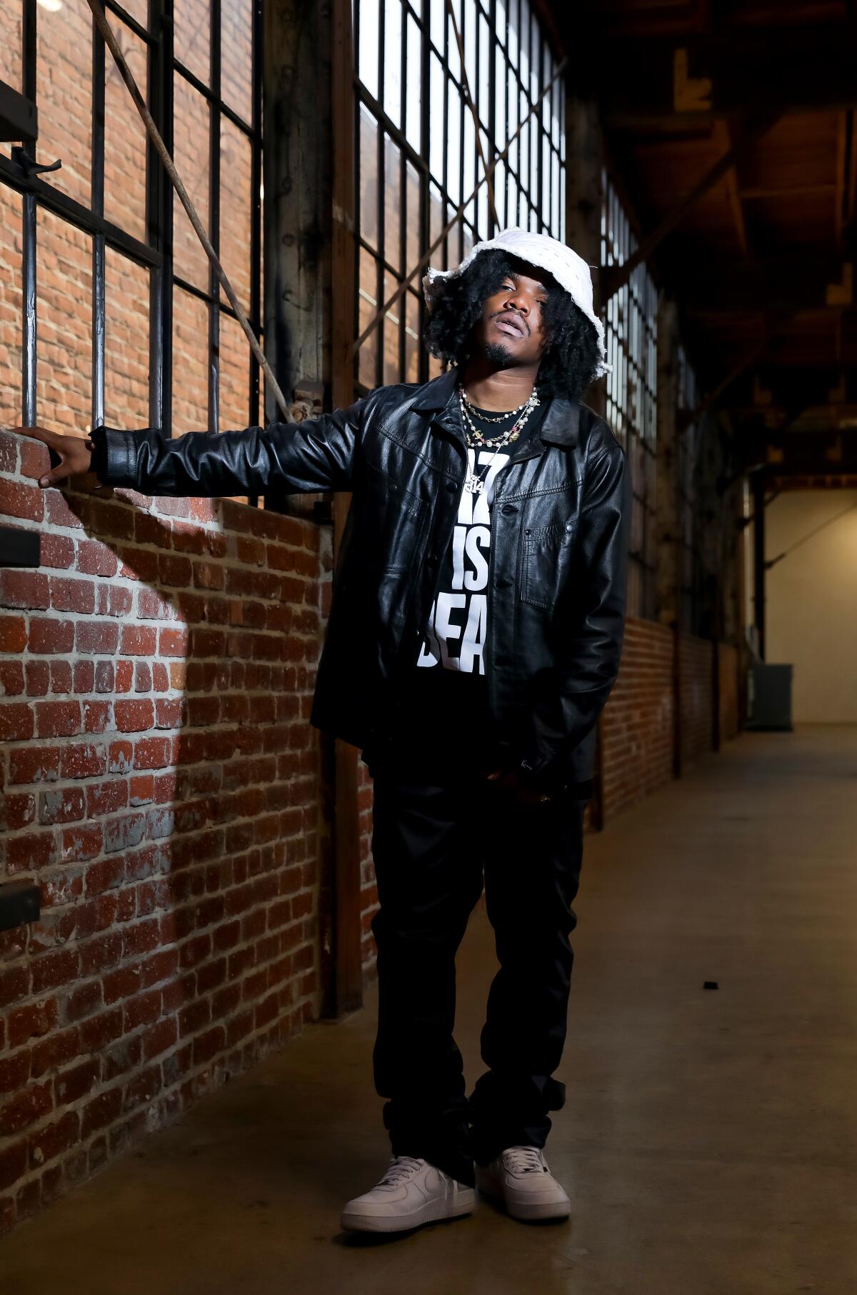 A rapper in a white bucket hat stands against a brick wall