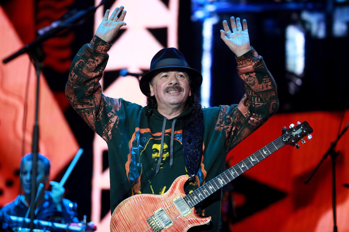A guitarist in a black hat and colorful shirt holds his hands up onstage