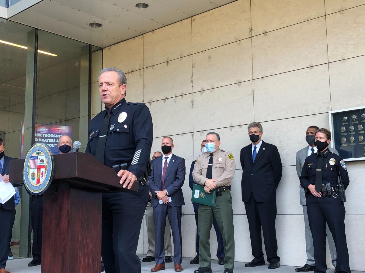 LAPD Chief Michel Moore speaks at a lectern with other masked law enforcement leaders standing behind him outside LAPD HQ