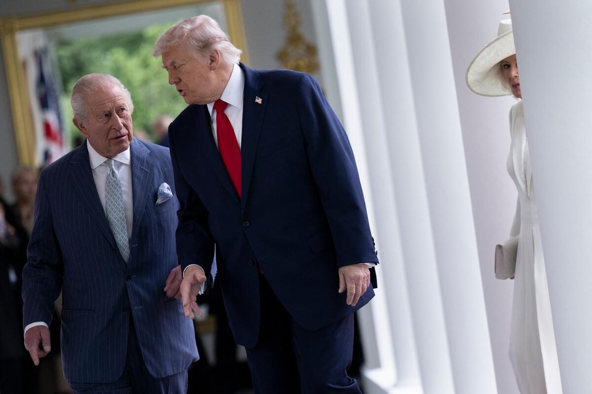 Two men in dark suits, one with a pale blue tie, the other red, walking near a partially visible woman in white hat and dress