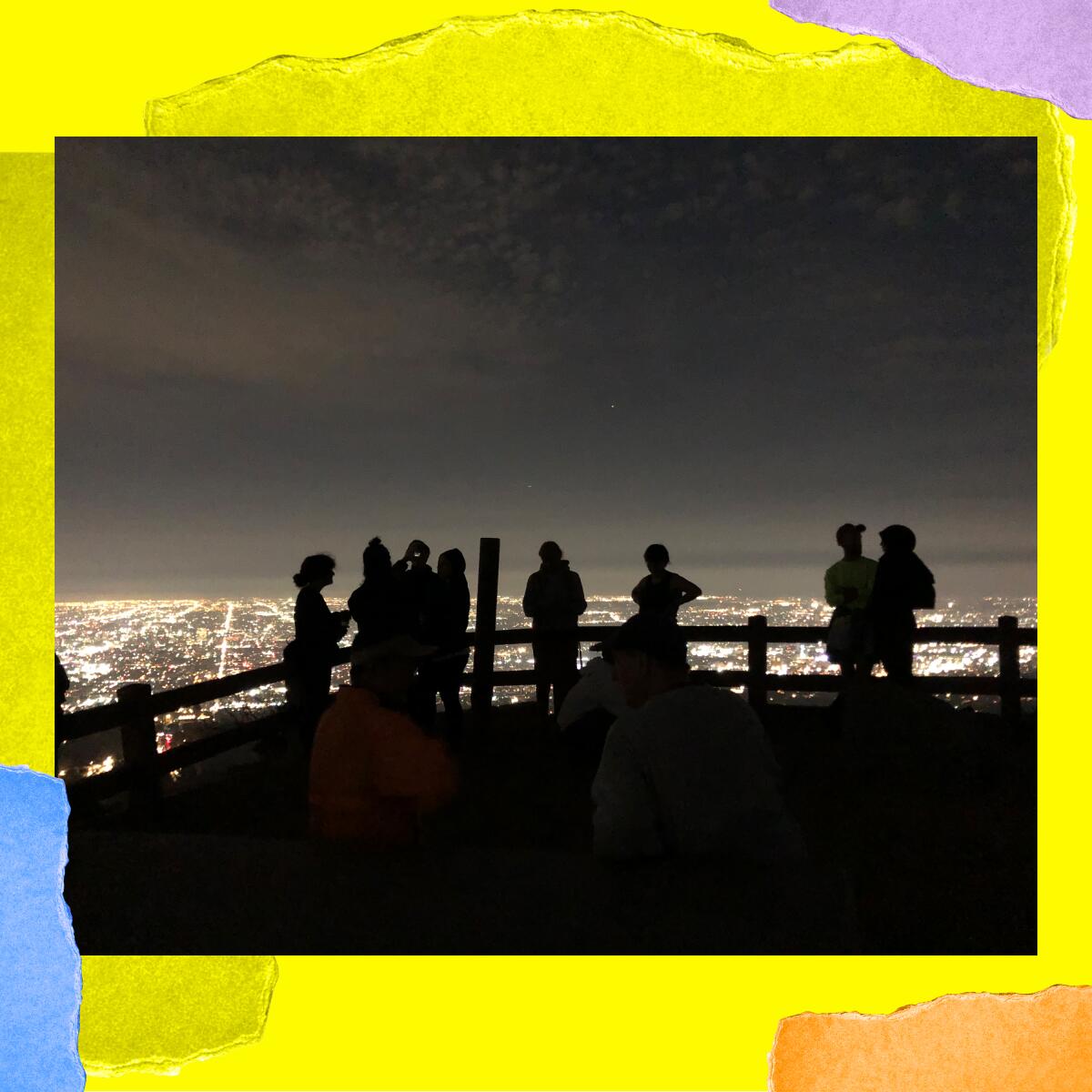 People standing against a split-rail fence are silhouetted by city lights that stretch to the horizon in a valley below.