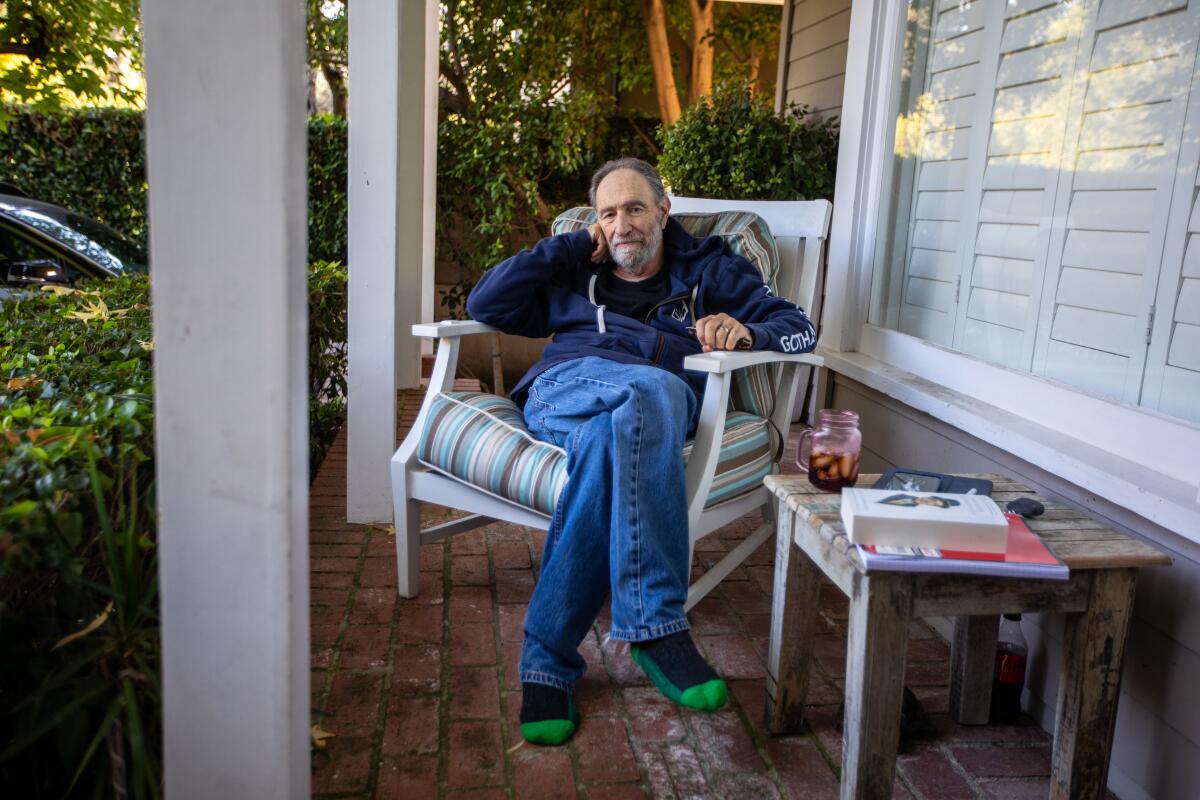 An older man sits with his legs crossed in a chair on a porch.