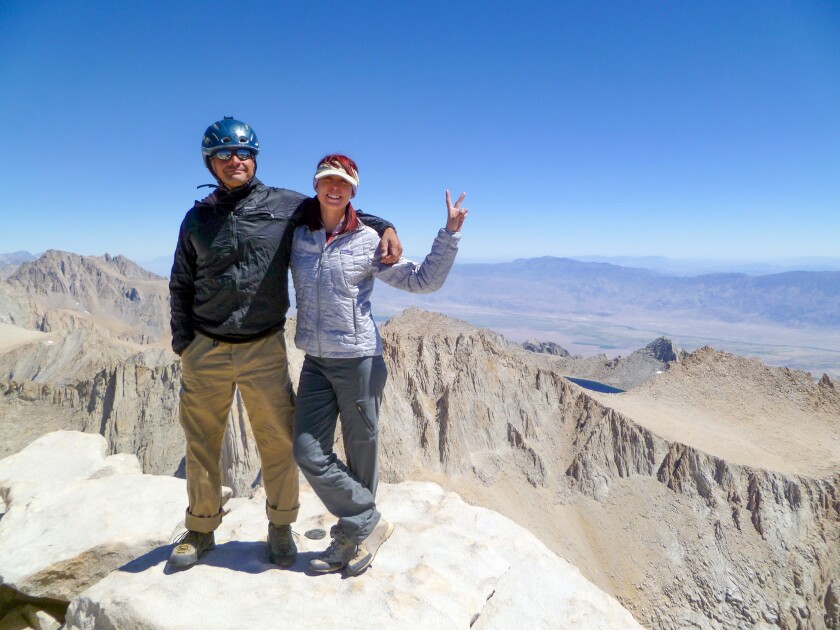 A man and a woman stand on a mountaintop, his arm around her shoulder. 