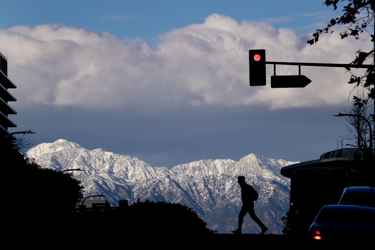 A pedestrian crossing a street in downtown Los Angeles is silhouetted against the snow-capped San Gabriel Mountains.