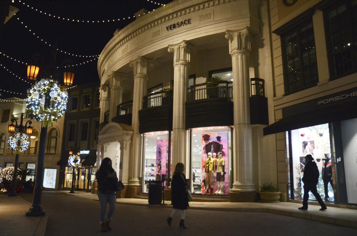 People walk near the Versace boutique on Rodeo Drive, decorated for the holidays amid the coronavirus pandemic, Monday, Nov. 30, 2020, in Beverly Hills, Calif. The three-week "safer at home" order began on Monday. Indoor retail businesses, which make much of their profits during the Thanksgiving and Christmas holiday seasons, are allowed to remain open but with just 20% of capacity, including nail salons and other personal care services. (AP Photo/Pamela Hassell)