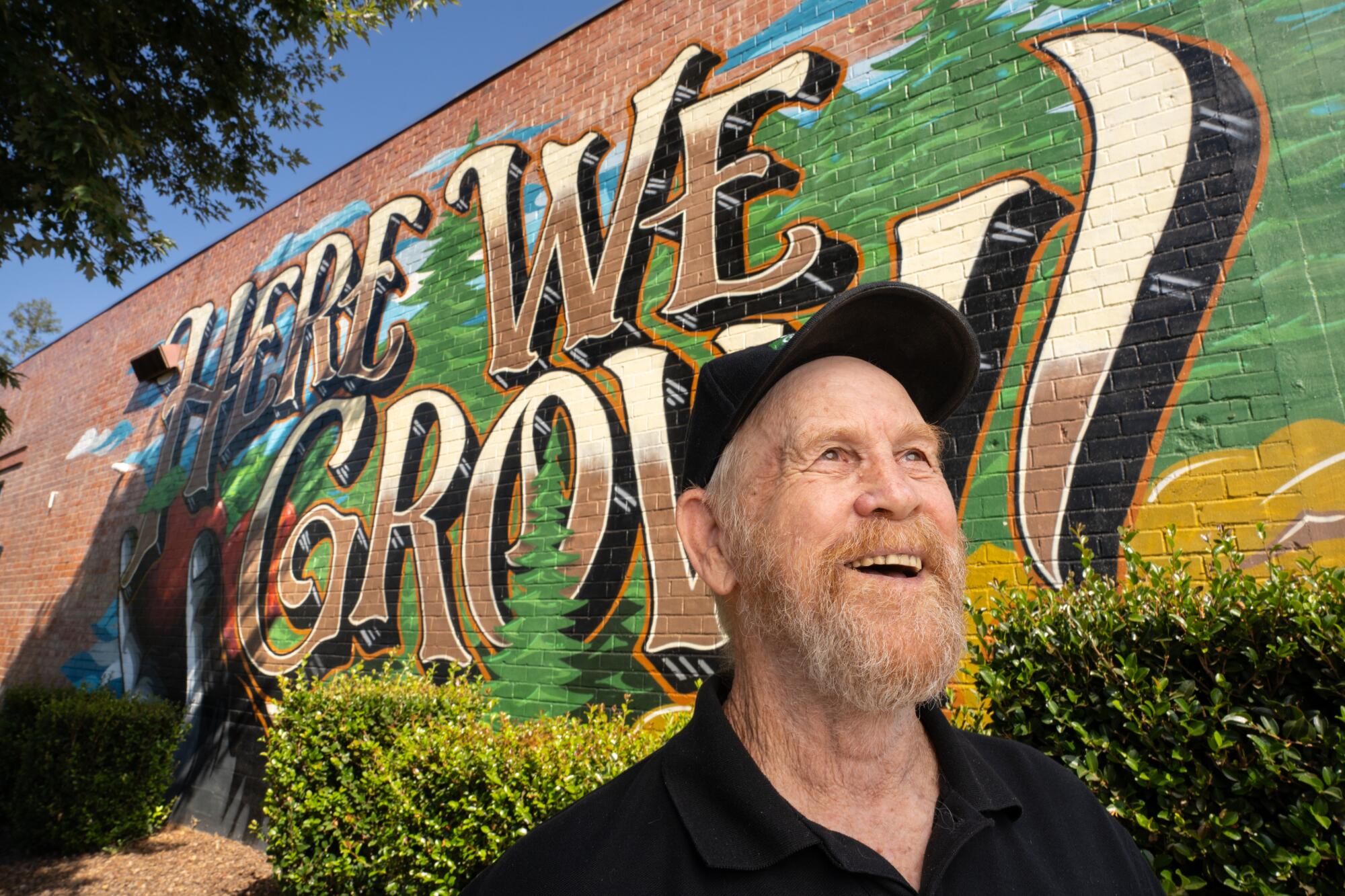 A bearded man smiles in front of a mural.
