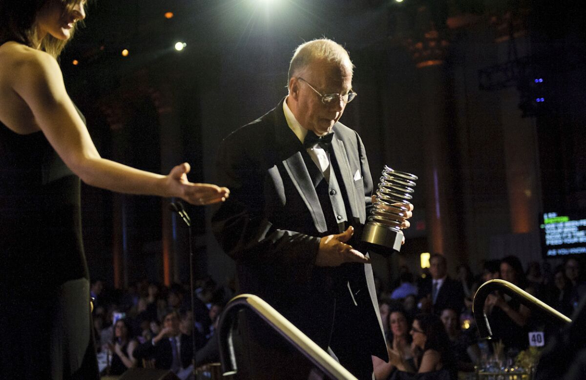 A man in a tuxedo walks offstage holding a cylindrical-shaped award