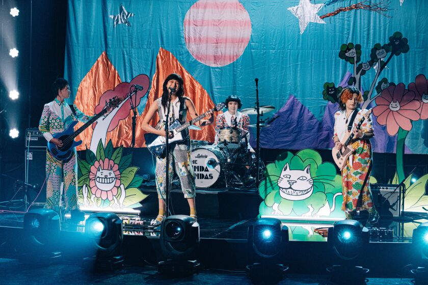 The four members of rock band the Linda Lindas playing guitar and drums on a stage
