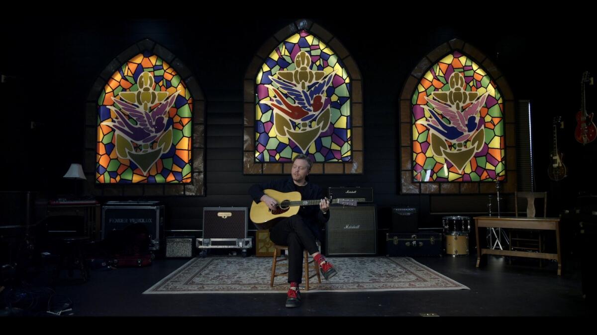 man plays guitar with three large stained glass windows behind him 