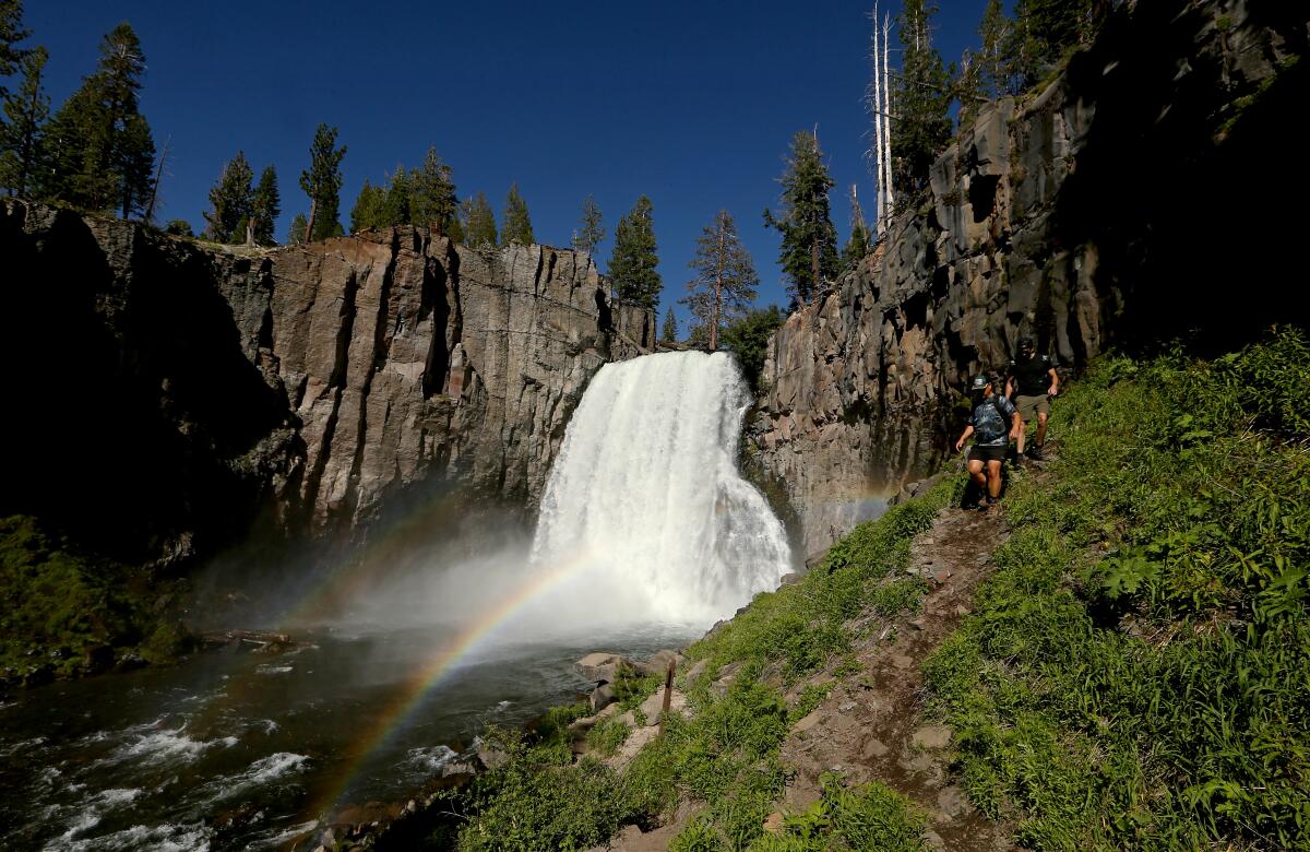 People walk down a steep trail next to a large waterfall between rocky cliffs. A rainbow is visible in the foreground.