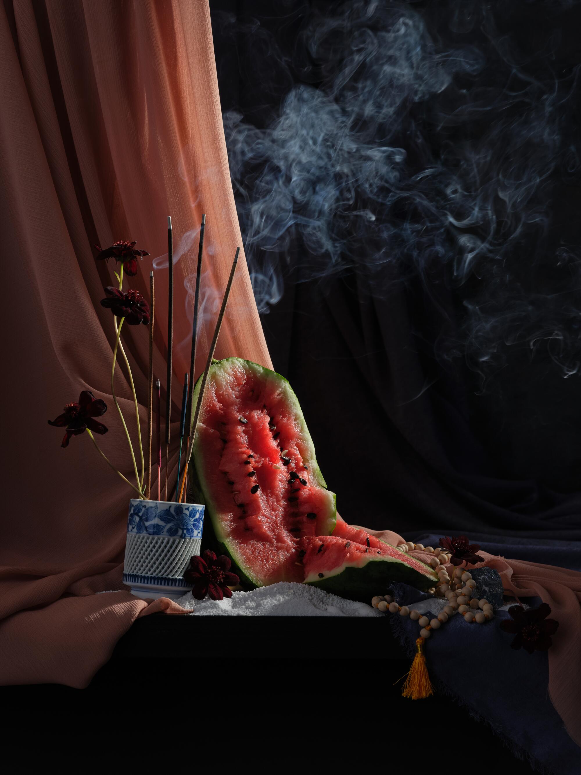 a ceramic jar filled with incense sticks and tall flowers next to an open watermelon leaning against a coral curtain