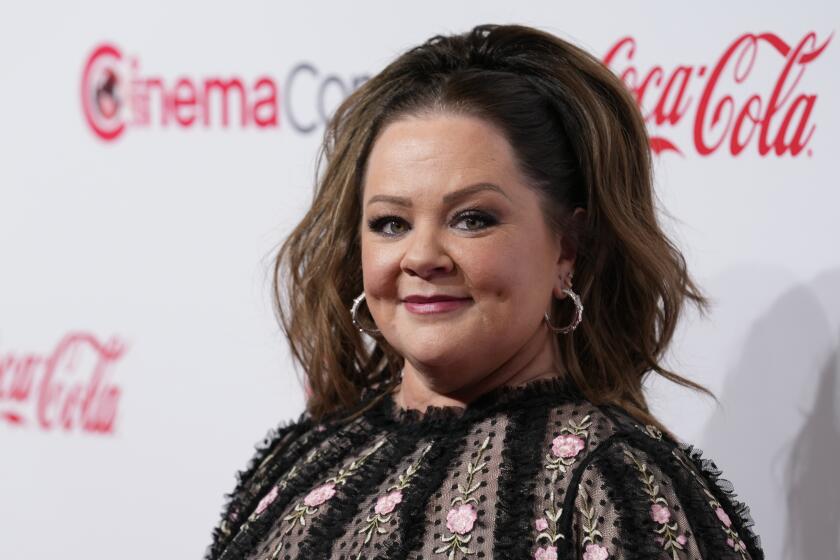 Melissa McCarthy is posing and smiling while wearing a black flowered dress with her hair up. 