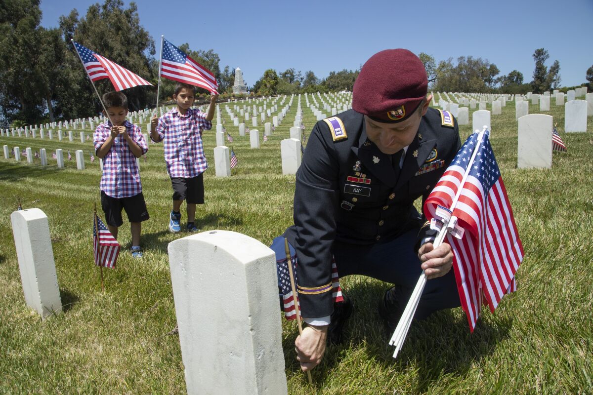 A man in uniform kneels to put a miniature flag in the ground at a gravestone. Behind him, two young boys hold flags.