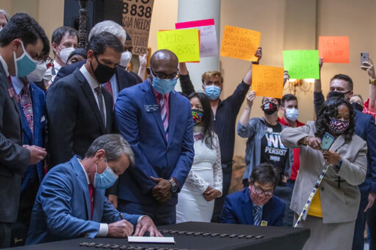 Gov. Brian Kemp, lower left, signs HB 426, hate-crimes legislation, into law as demonstrators hold signs asking him to veto HB 838 in the North Wing of the Georgia State Capitol building Friday, June 26, 2020. Kemp signed into law House Bill 426 which would impose additional penalties for crimes motivated by the victim’s race, color, religion, national origin, sexual orientation, gender or disability. (Alyssa Pointer/Atlanta Journal-Constitution via AP)